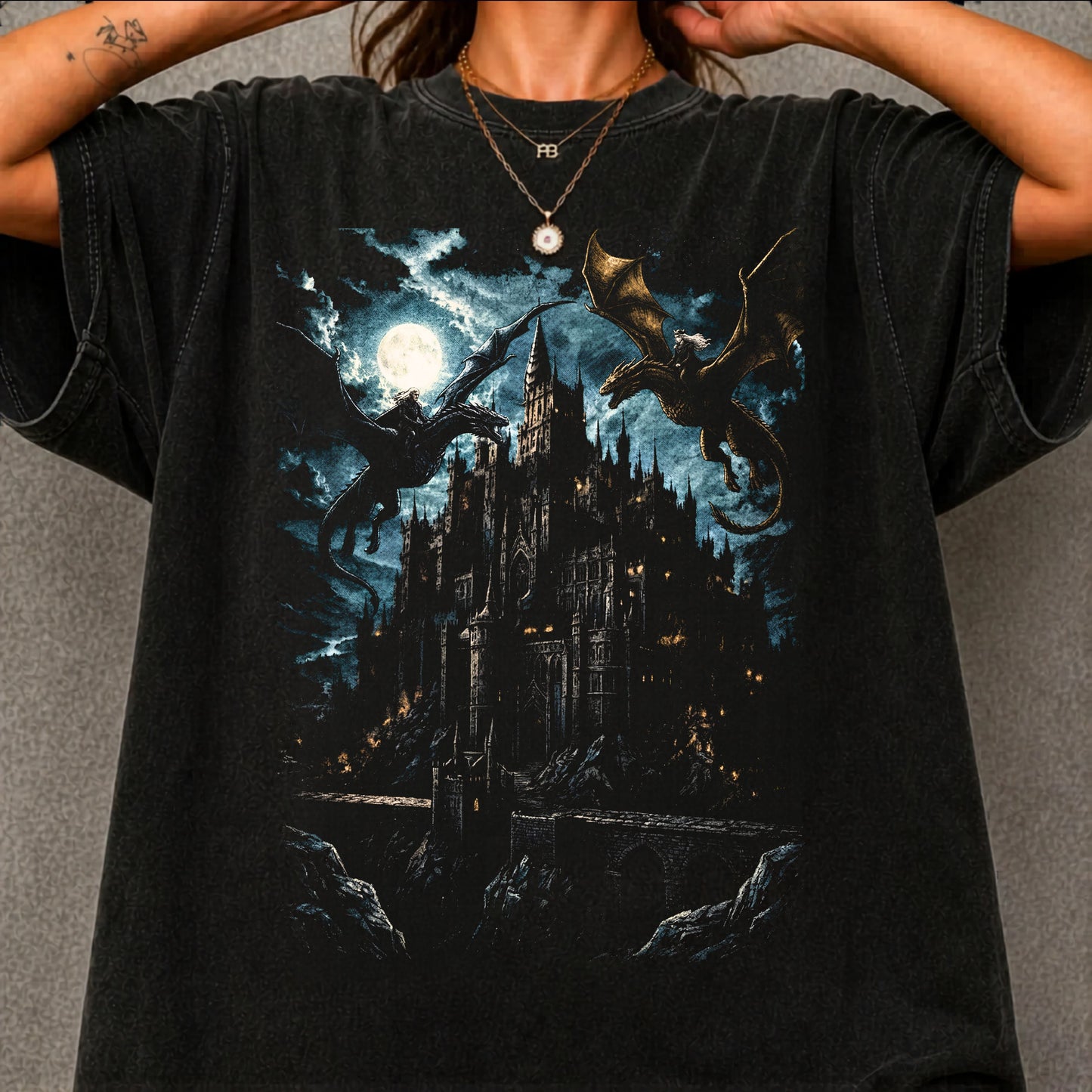 A person wearing a black graphic t-shirt featuring a dark fantasy illustration of a gothic castle under a full moon with dragons, set against a gray textured background.