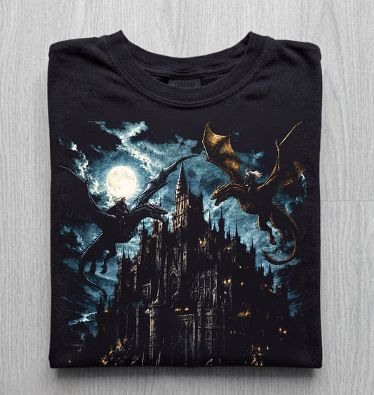 Folded black graphic tee featuring a gothic castle under a full moon with two dragons soaring through a dark indigo night sky, displayed on a light gray wood-textured background.
