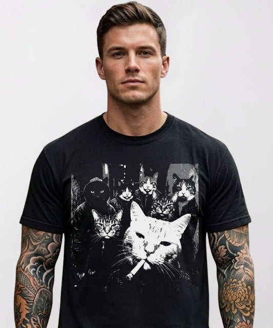 Black heavyweight graphic t-shirt featuring a moody black-and-white photo-style print of six tough cats posed like a crime gang, with a white boss cat holding a cigarette in the foreground, worn by a tattooed male model.