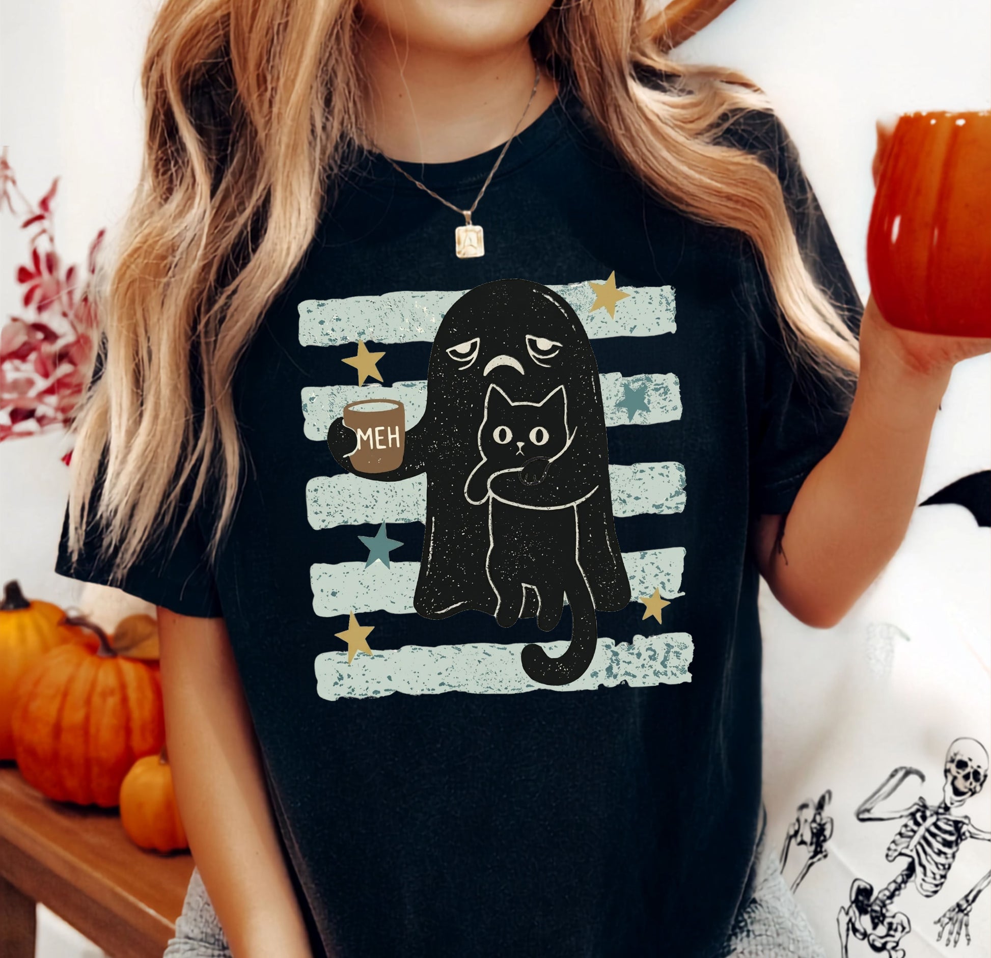 A woman wearing a black graphic t-shirt with a cartoon cat and ghost design, holding an orange mug in a cozy, autumn-themed setting with pumpkins and a skeleton tote bag.
