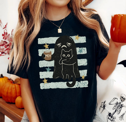 A woman wearing a black graphic t-shirt with a cartoon cat and ghost design, holding an orange mug in a cozy, autumn-themed setting with pumpkins and a skeleton tote bag.
