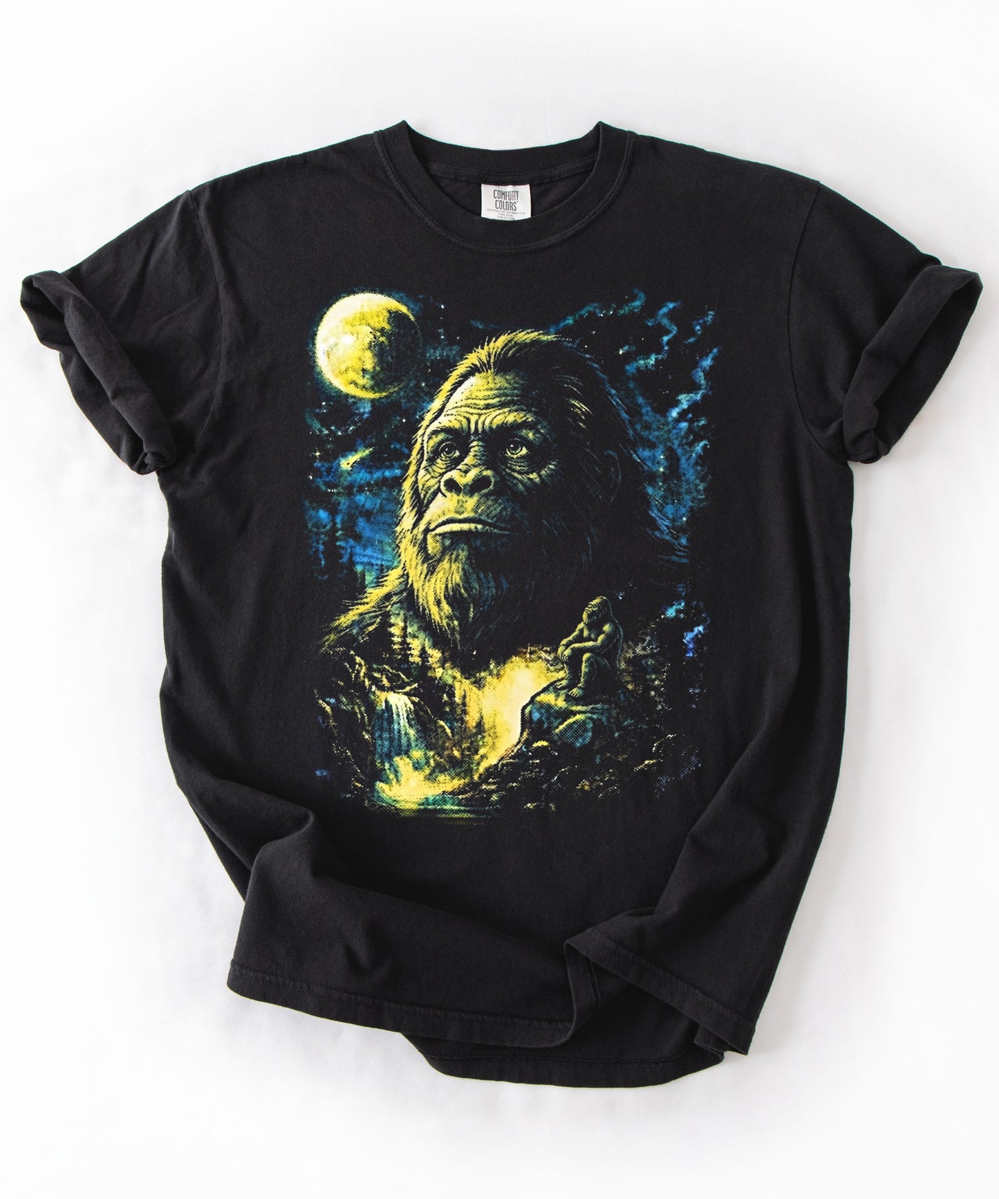A black short-sleeve t-shirt with rolled cuffs, featuring a graphic print of Bigfoot under a full moon in a blue and yellow night sky, laid flat on a white background.