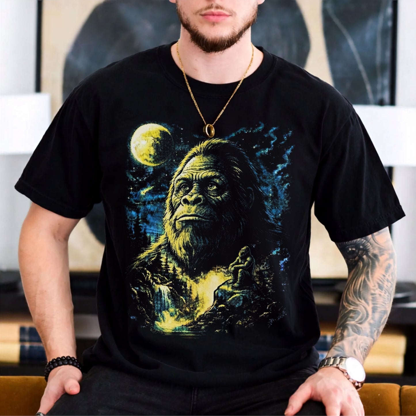 A man wearing a black short-sleeve graphic t-shirt featuring a large print of Bigfoot under a full moon, with a blurred indoor background.