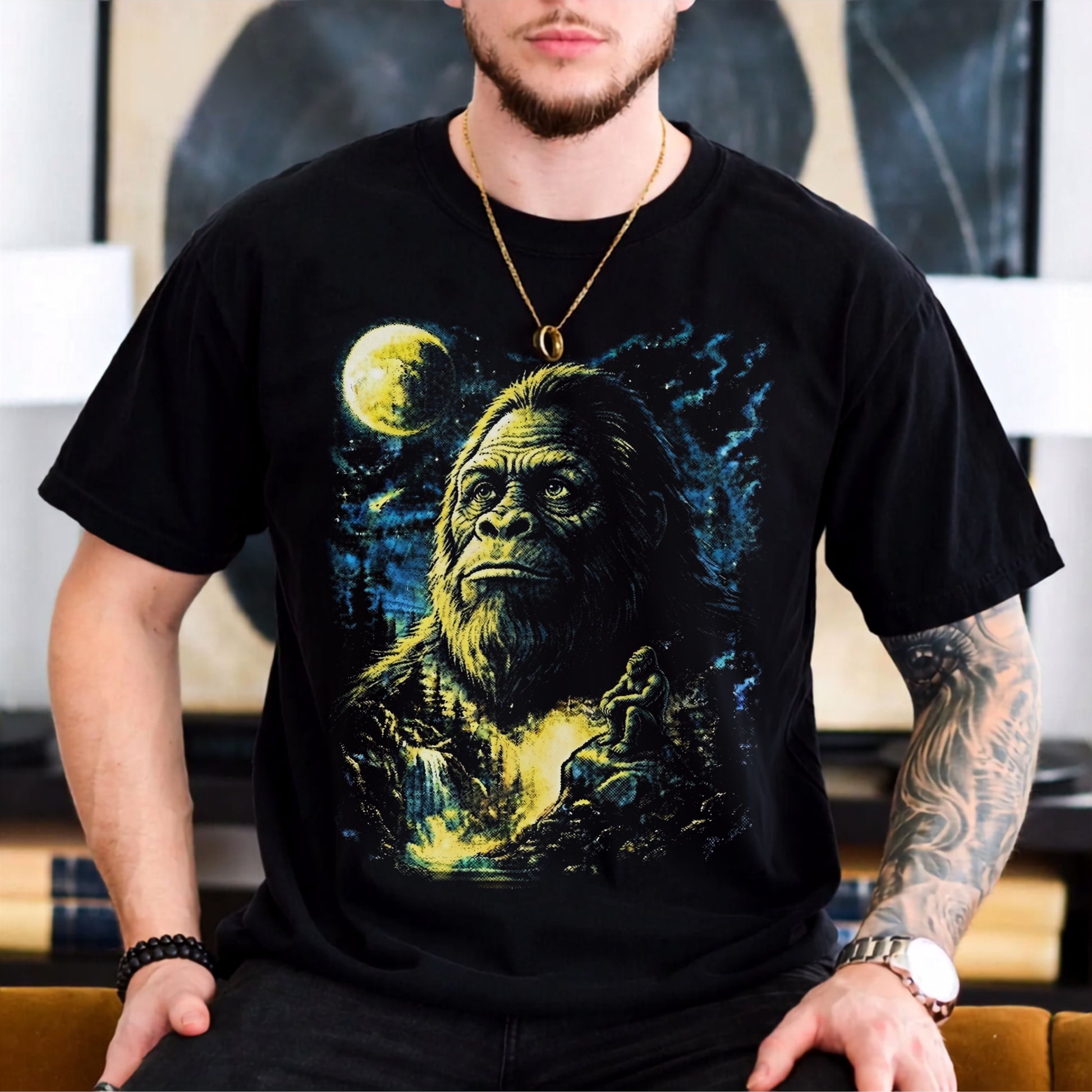 A man wearing a black short-sleeve graphic t-shirt featuring a large print of Bigfoot under a full moon, with a blurred indoor background.