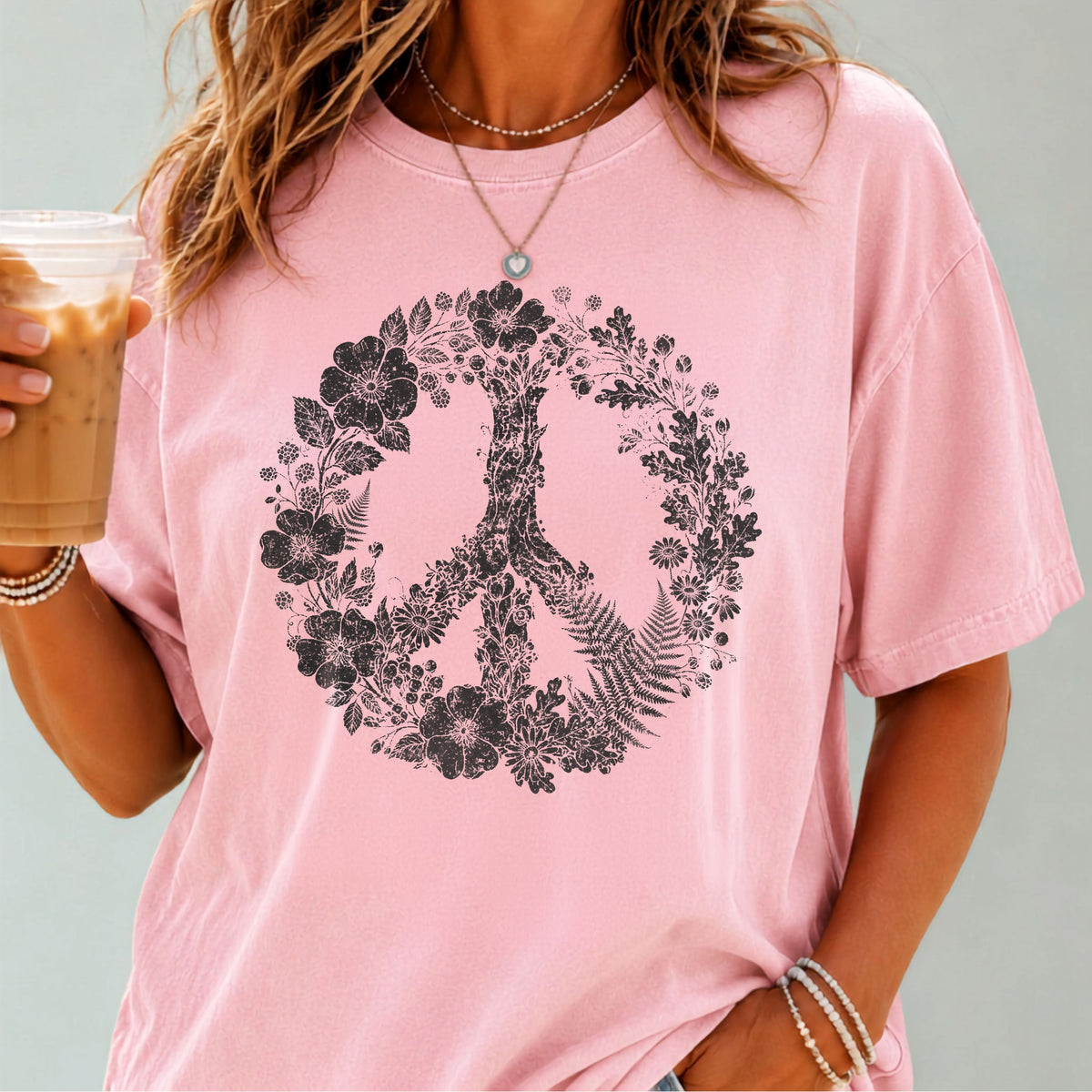 A woman wearing a Blossom pink short-sleeve t-shirt with a black peace sign graphic filled with flowers and leaves. She is holding an iced coffee and wearing a necklace and bracelets against a solid gray background.