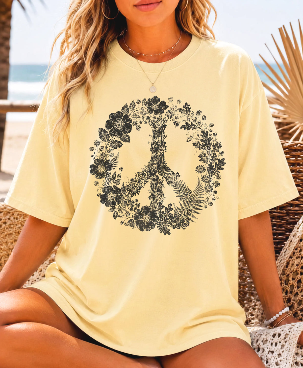A woman wearing a Butter yellow oversized T-shirt with a large black floral peace sign graphic on the front, seated on a beach chair with a beach background.