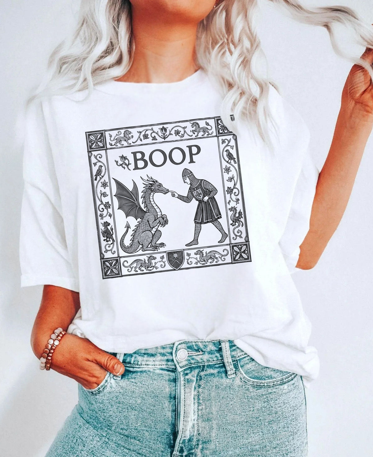 oop Dragon T-Shirt in White — medieval manuscript-style graphic of a knight booping a dragon, worn by a model with light-wash jeans