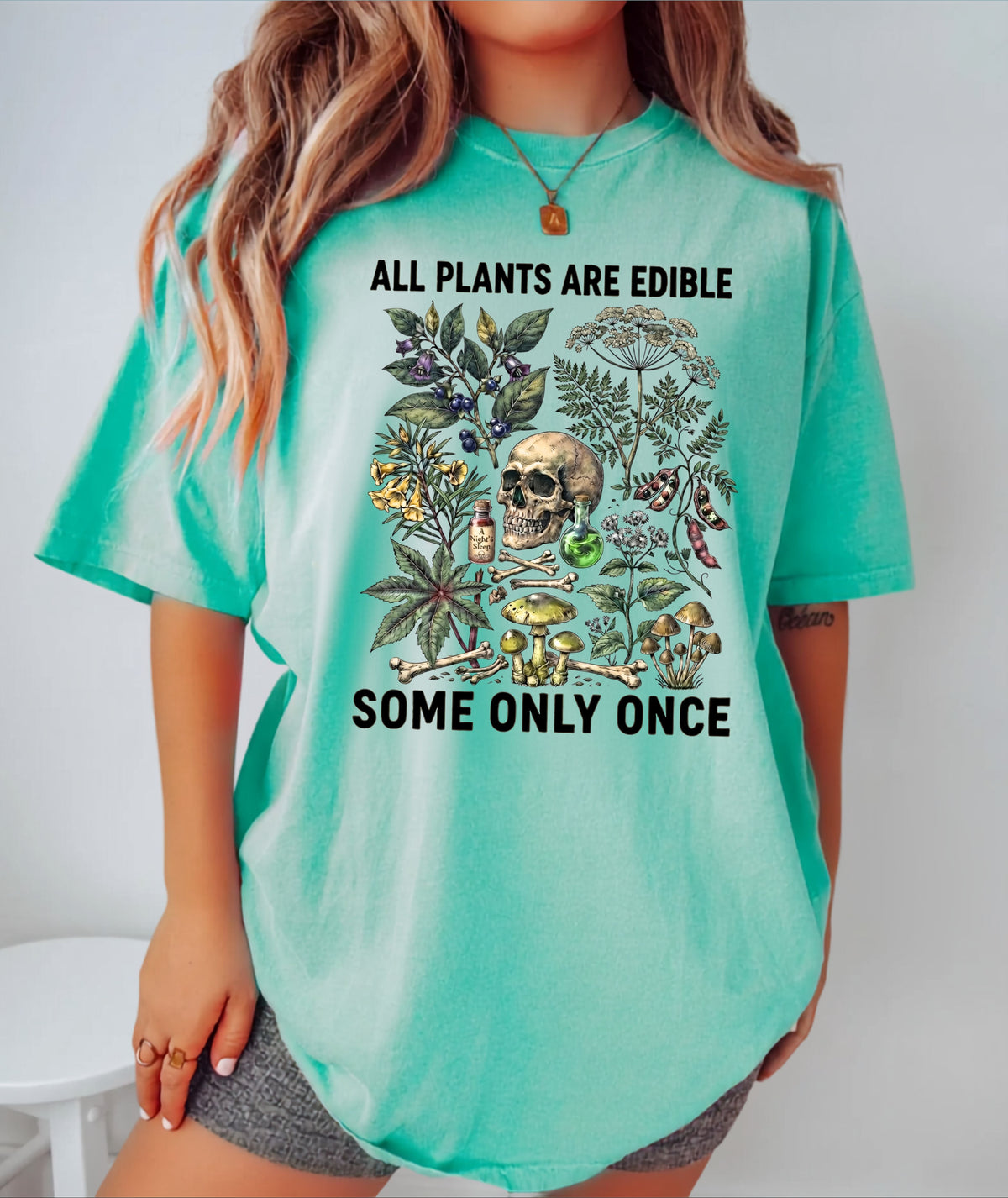 A woman wearing an Island Reef  oversized graphic t-shirt with the text 'ALL PLANTS ARE EDIBLE SOME ONLY ONCE' and a botanical skull print, paired with gray shorts.