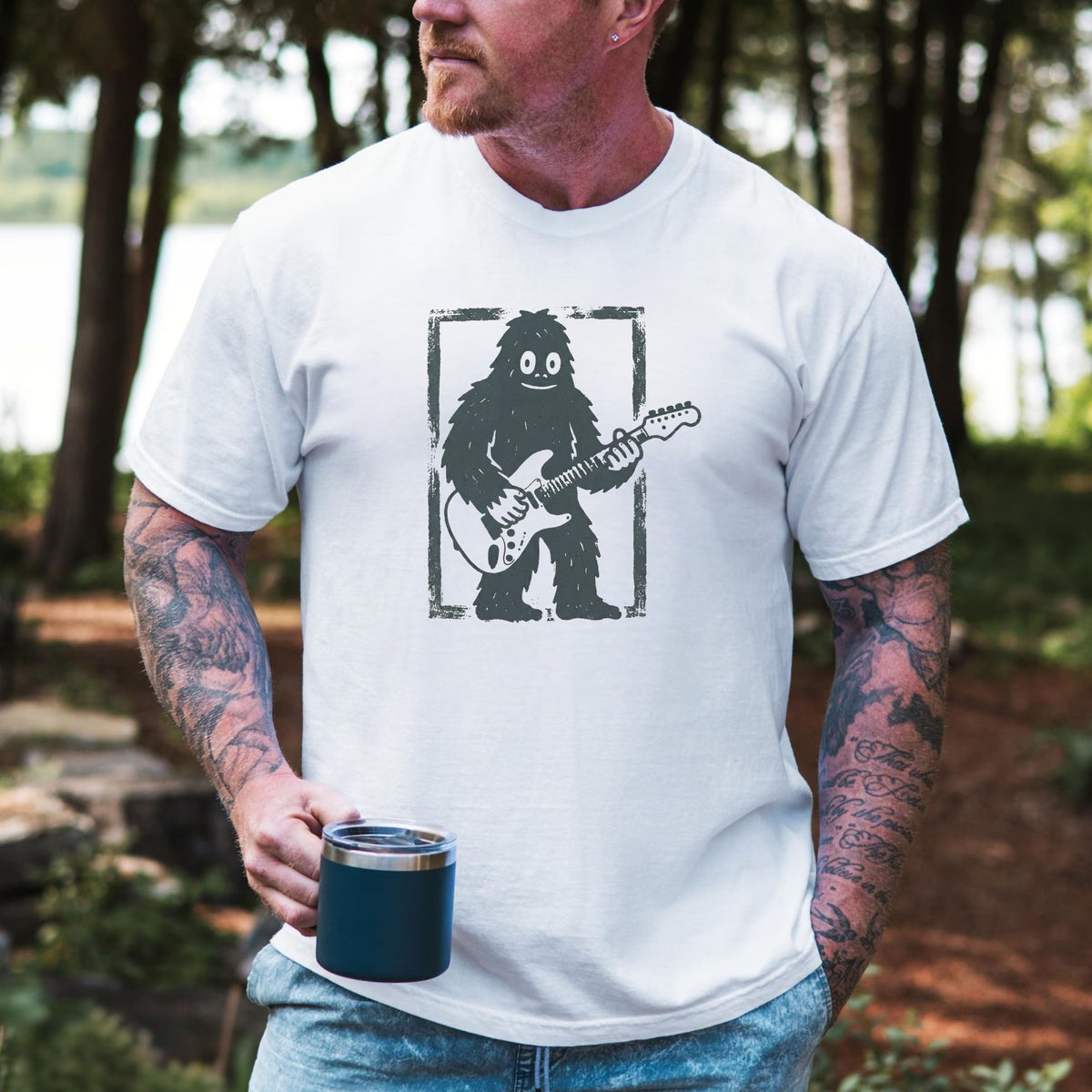 A man wearing a white graphic t-shirt with a black and gray 'Bigfoot playing guitar' design, holding a dark blue insulated tumbler in an outdoor forest setting.
