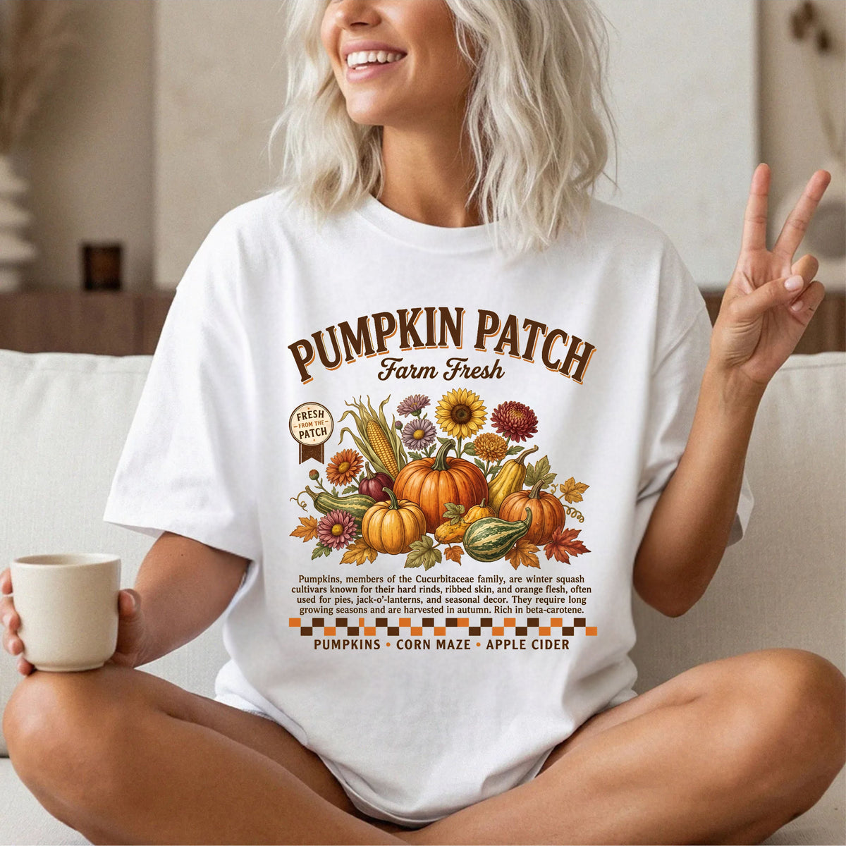 A woman sitting cross-legged indoors wearing a white graphic T-shirt with a colorful autumn 'Pumpkin Patch Farm Fresh' print. She is holding a mug in one hand and making a peace sign with the other, with a couch and home interior in the background.