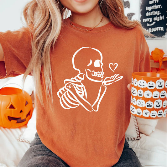A woman wearing a Yam orange crewneck t-shirt with a white line-art graphic of a skeleton Blowing a heart kiss. She is holding an orange pumpkin bucket and a matching pumpkin-patterned mug, with Halloween decorations in the background.