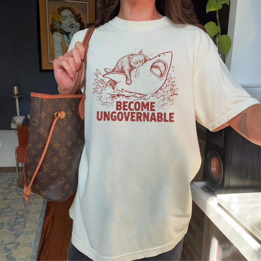 A person wearing an oversized Ivory t-shirt with a red graphic of a cat on a Shark and the text 'BECOME UNGOVERNABLE'. The person is holding a brown Louis Vuitton monogram handbag in a home environment.