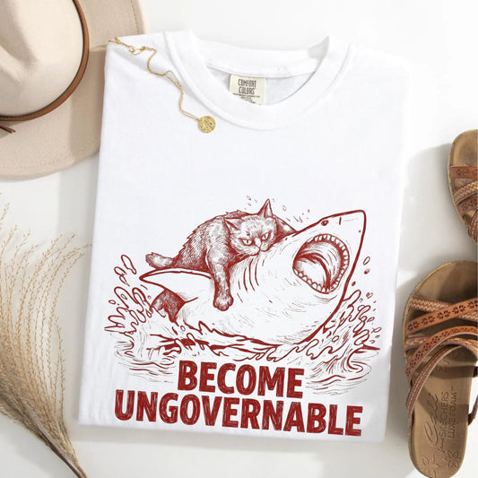 A white short-sleeve graphic T-shirt laid flat on a light surface, featuring a red sketch of a cat riding a shark with the text 'BECOME UNGOVERNABLE' below. A beige hat, dried grass, and brown sandals are styled around it.