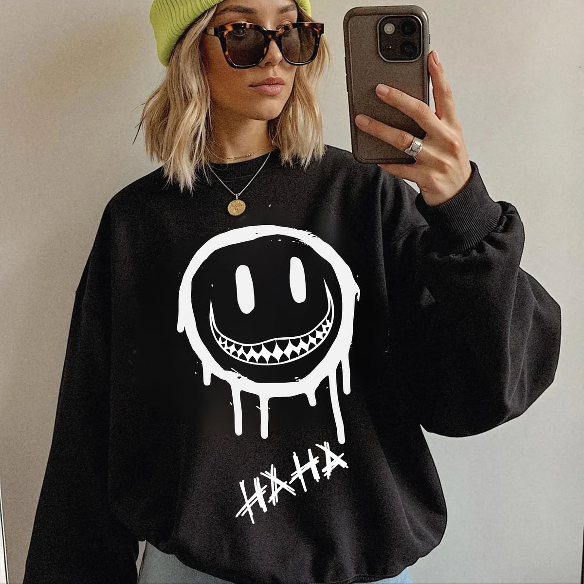 A woman takes a mirror selfie wearing a black sweatshirt with a large white dripping smiley face graphic and the word 'HAHA' below it. She is also wearing a lime green beanie, sunglasses, and a gold necklace.