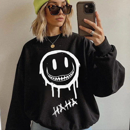 A woman takes a mirror selfie wearing a black sweatshirt with a large white dripping smiley face graphic and the word 'HAHA' below it. She is also wearing a lime green beanie, sunglasses, and a gold necklace.