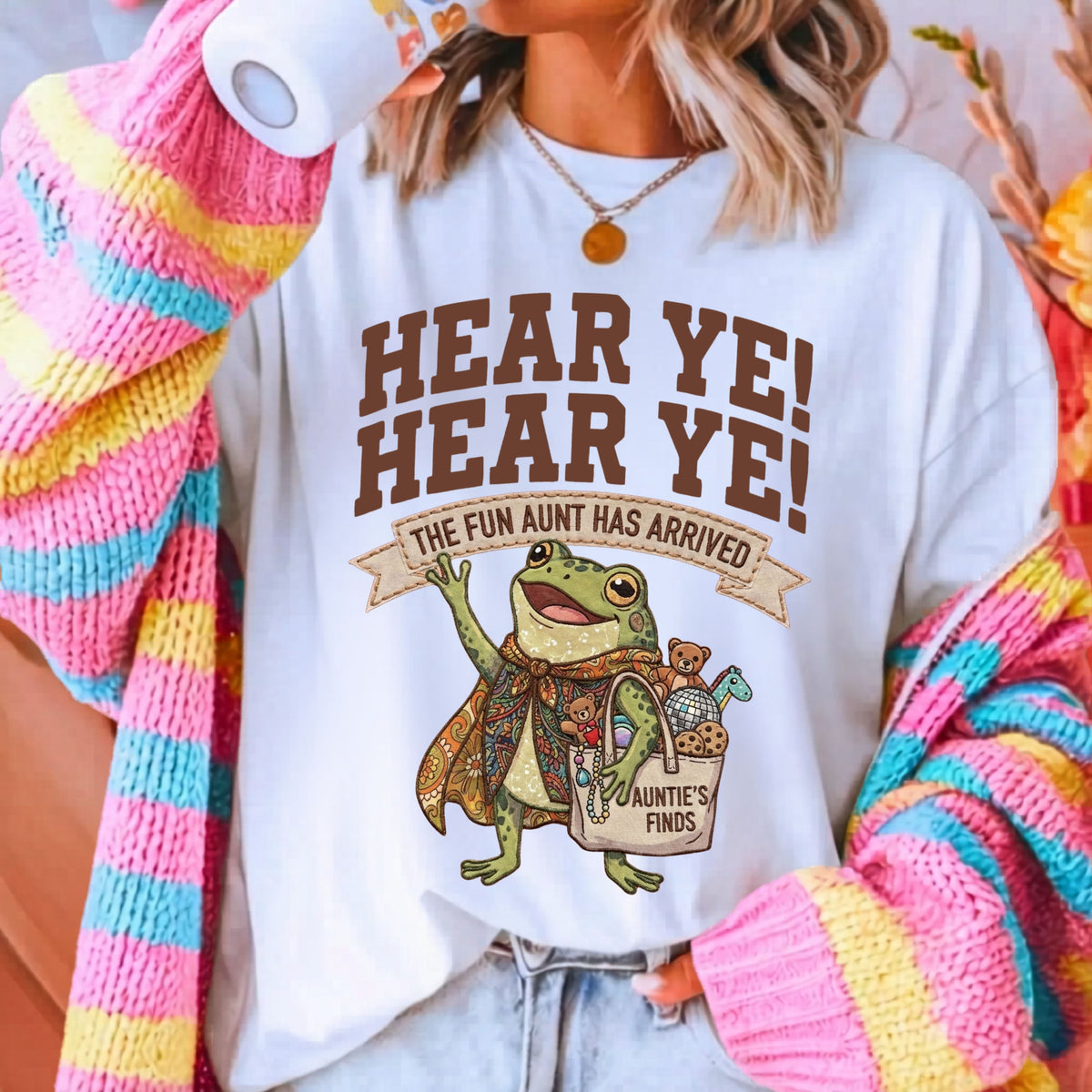 A woman wearing a white graphic t-shirt with a brown frog and text that reads 'HEAR YE! HEAR YE! THE FUN AUNT HAS ARRIVED'. She is also wearing a colorful pink, yellow, and blue striped knit cardigan and light wash jeans.