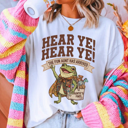A woman wearing a white graphic t-shirt with a brown frog and text that reads 'HEAR YE! HEAR YE! THE FUN AUNT HAS ARRIVED'. She is also wearing a colorful pink, yellow, and blue striped knit cardigan and light wash jeans.