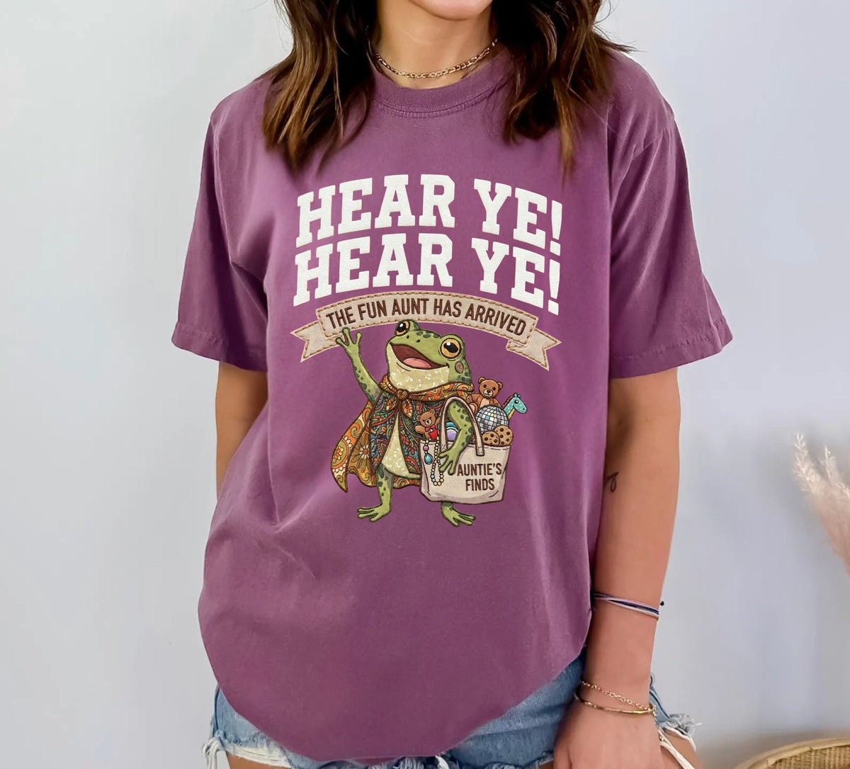 A woman wearing a Berry purple graphic t-shirt with a cartoon frog and the text 'HEAR YE! HEAR YE! THE FUN AUNT HAS ARRIVED!'.