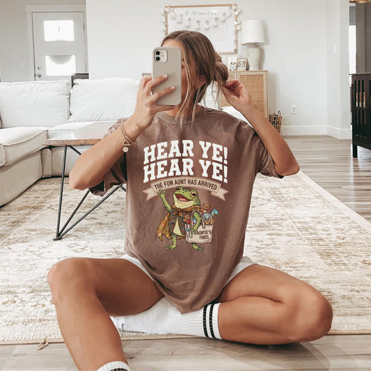 A woman sitting cross-legged on a light-colored rug in a living room, taking a mirror selfie while wearing a brown graphic t-shirt with a cartoon frog and the text 'HEAR YE! HEAR YE! THE FUN Aunt HAS ARRIVED'.