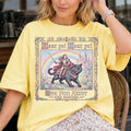 A woman wearing a Butter yellow graphic t-shirt with a vintage-style illustration of a woman riding a black cat under a rainbow and the text 'Hear ye! Hear ye! The Fun Aunt Is Arrived'.