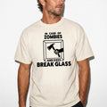 A man wearing a light beige crewneck t-shirt with a black graphic print that reads 'IN CASE OF ZOMBIES AND EXES BREAK GLASS' with an illustration of a hand holding an axe. The photo is taken against a white wall with yellow borders at the top and bottom.