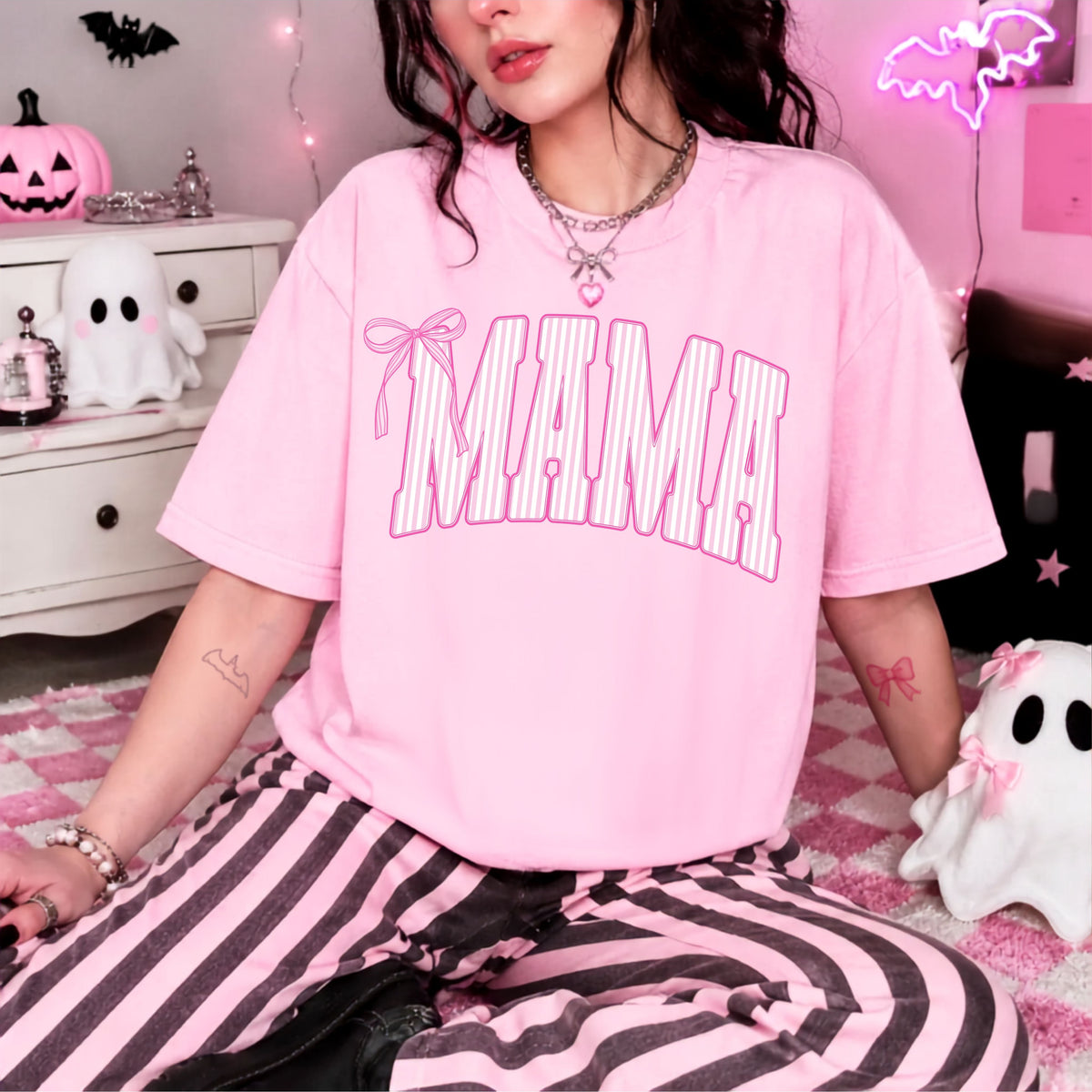 A person wearing an oversized light pink T-shirt with the word "MAMA" in large arched pink letters outlined with white stripes, paired with pink-and-black striped pants, seated in a pink Halloween-themed room.