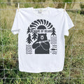 A white short-sleeve t-shirt with a black graphic print of a bear, surrounded by trees, waves, and a fish, with the text 'BEWARE OF BEARS' across the chest. The shirt is hanging from a wire fence in an outdoor grassy field.