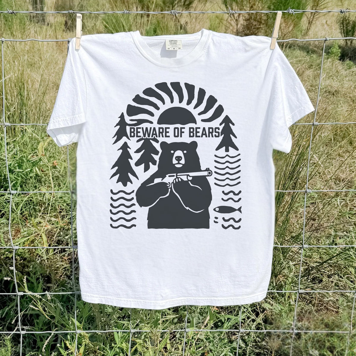 A white short-sleeve t-shirt with a black graphic print of a bear, surrounded by trees, waves, and a fish, with the text 'BEWARE OF BEARS' across the chest. The shirt is hanging from a wire fence in an outdoor grassy field.