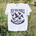 White Comfort Colors Fly You Fools tee with celestial birds, mountains, and pine trees outdoors
