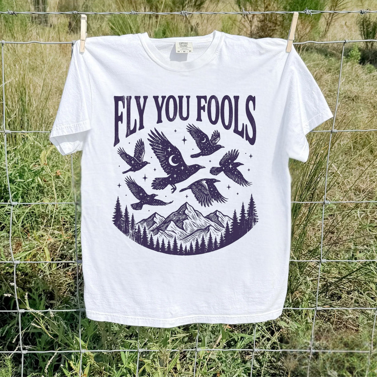 White Comfort Colors Fly You Fools tee with celestial birds, mountains, and pine trees outdoors