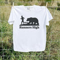 A white short-sleeve t-shirt hanging on a wire fence outdoors, featuring a black graphic of a runner and a bear above the text 'Runners High'.