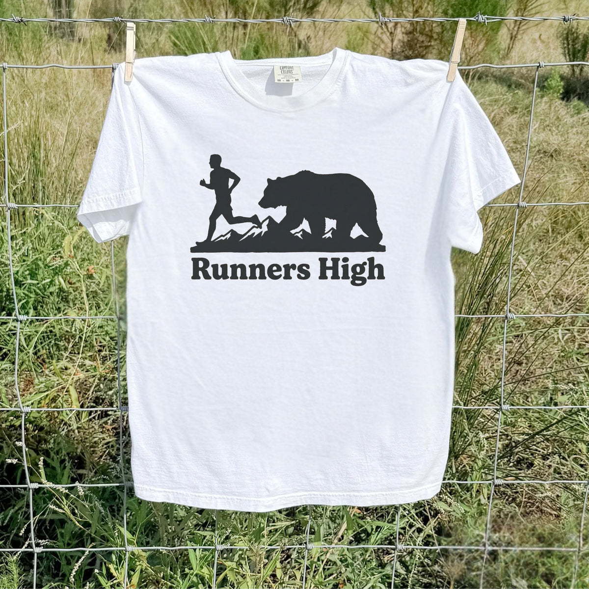 A white short-sleeve t-shirt hanging on a wire fence outdoors, featuring a black graphic of a runner and a bear above the text 'Runners High'.