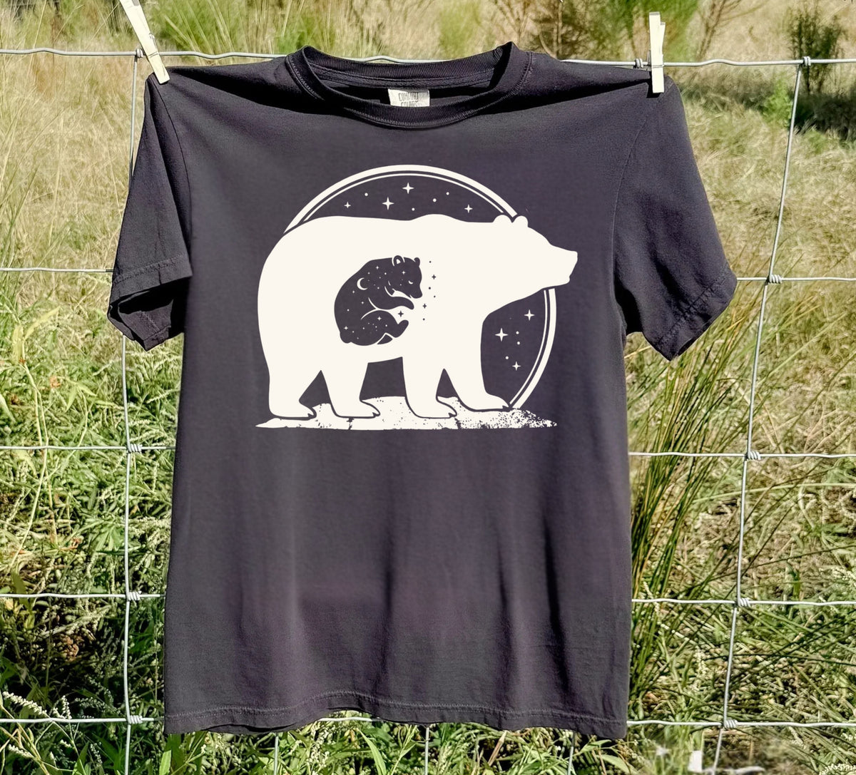 A Black colored short-sleeve t-shirt with a black graphic of a bear silhouette containing a baby moon and stars, laid flat on a white background.