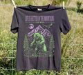 A black graphic t-shirt hanging on a wire fence outdoors. The shirt features a green and purple graphic of a grizzly bear in a mountain forest, with the text 'LIFE IS BETTER IN THE MOUNTAINS' and 'LIVE WILD.