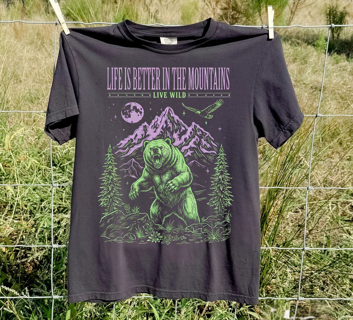 A black graphic t-shirt hanging on a wire fence outdoors. The shirt features a green and purple graphic of a grizzly bear in a mountain forest, with the text 'LIFE IS BETTER IN THE MOUNTAINS' and 'LIVE WILD.