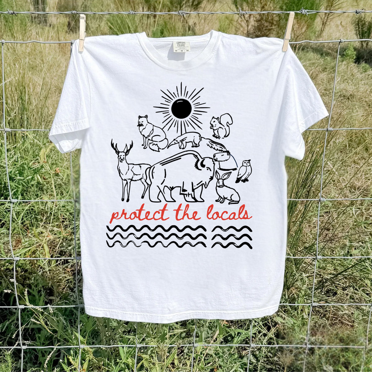 A white short-sleeve graphic T-shirt hanging on a wire fence outdoors. The shirt features a black line-art illustration of animals and a sun with the phrase 'protect the locals' in red script and black wavy lines below.