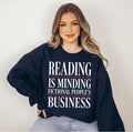 A woman wearing a navy blue sweatshirt with the text 'READING IS MINDING FICTIONAL PEOPLE'S BUSINESS' in white and red, paired with light blue jeans, set against a plain light gray background.
