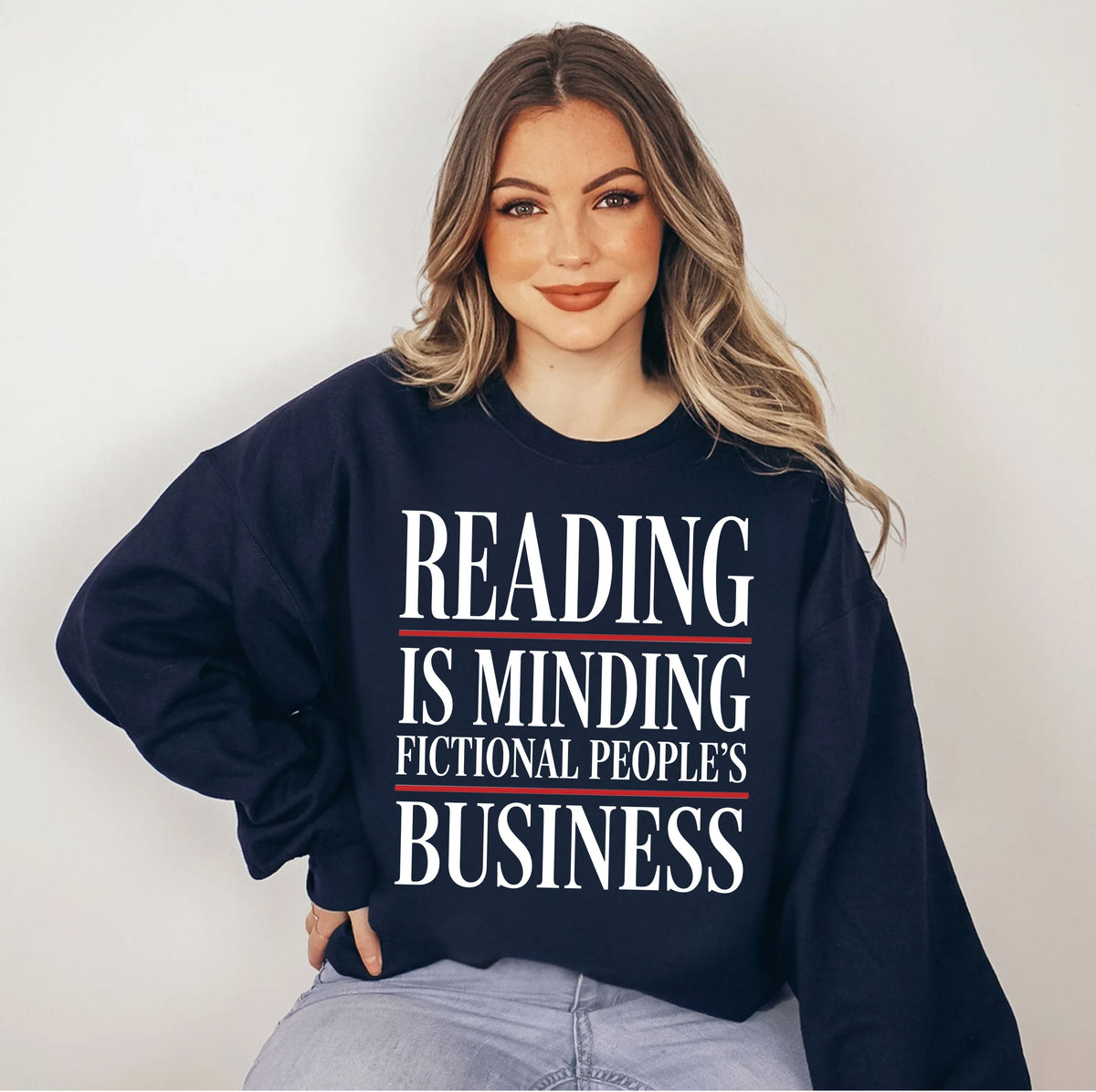 A woman wearing a navy blue sweatshirt with the text 'READING IS MINDING FICTIONAL PEOPLE'S BUSINESS' in white and red, paired with light blue jeans, set against a plain light gray background.