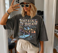 A woman takes a mirror selfie wearing an oversized charcoal pepper gray graphic t-shirt with the text 'LOST & OUT OF BREATH' and a mountain landscape print, paired with beige shorts and a gray cap.