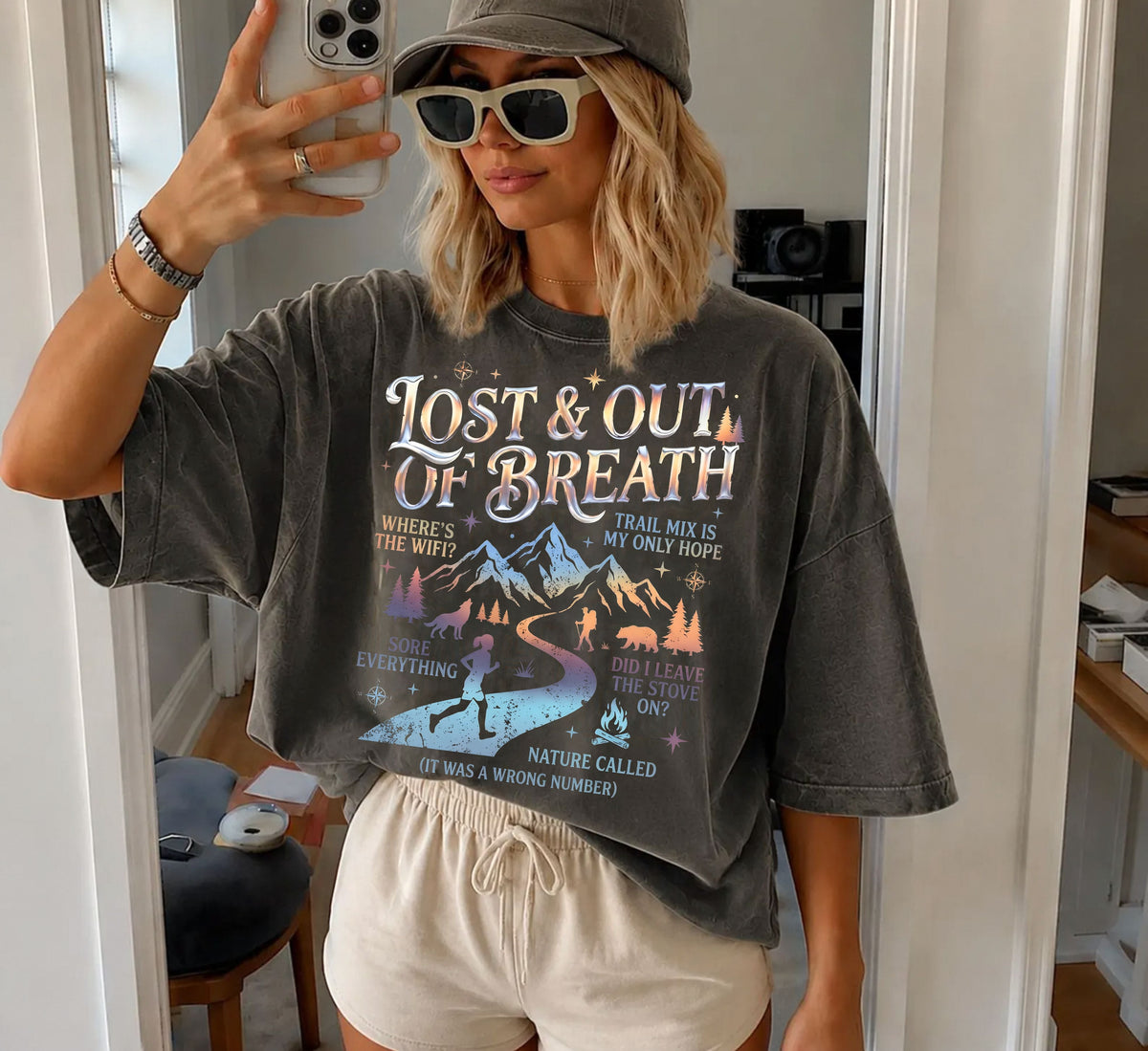 A woman takes a mirror selfie wearing an oversized charcoal pepper gray graphic t-shirt with the text 'LOST & OUT OF BREATH' and a mountain landscape print, paired with beige shorts and a gray cap.