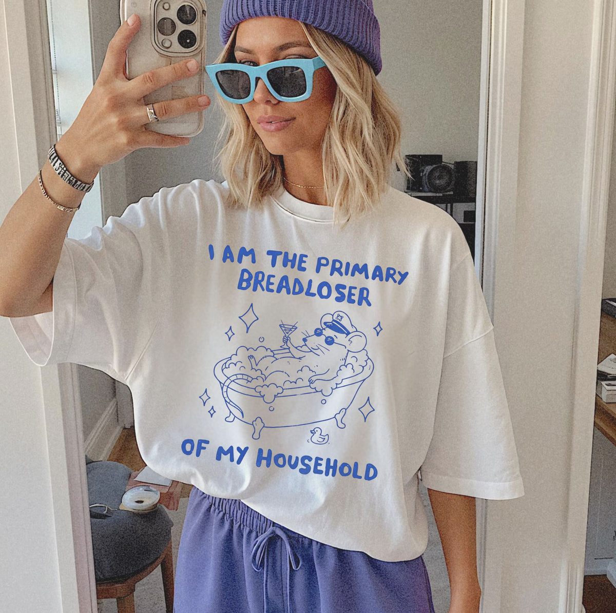 A woman taking a mirror selfie wearing an oversized white graphic t-shirt with blue text that reads 'I AM THE PRIMARY BREADLOSER OF MY HOUSEHOLD' and a cartoon drawing of a bubbly bathtub. She is also wearing a purple beanie, light blue sunglasses, and purple sweatpants.
