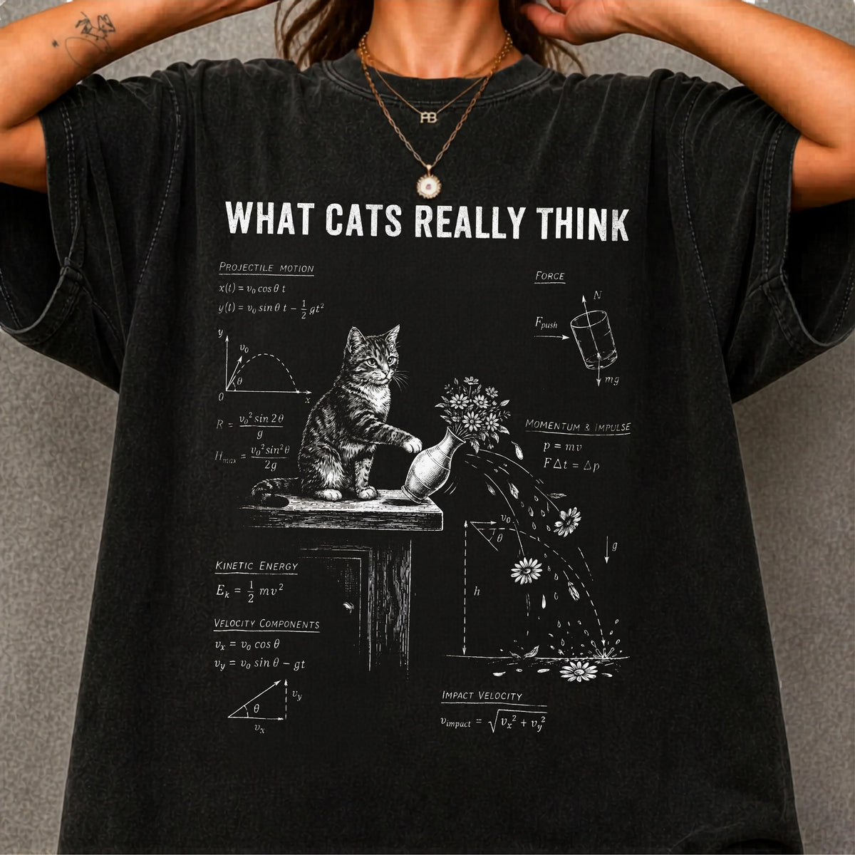 A folded black graphic T-shirt with the text 'WHAT CATS REALLY THINK' and a white cat illustration with diagrams, styled on a light background with a tan hat, gold necklace, and dried flowers.