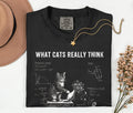 A folded black graphic T-shirt with the text 'WHAT CATS REALLY THINK' and a white cat illustration with diagrams, styled on a light background with a tan hat, gold necklace, and dried flowers.