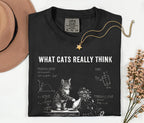 A folded black graphic T-shirt with the text 'WHAT CATS REALLY THINK' and a white cat illustration with diagrams, styled on a light background with a tan hat, gold necklace, and dried flowers.