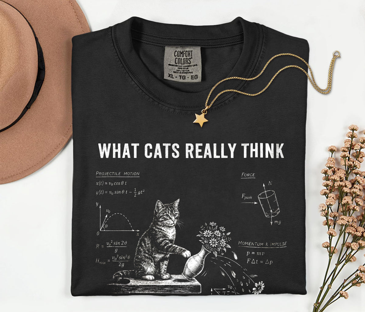 A folded black graphic T-shirt with the text 'WHAT CATS REALLY THINK' and a white cat illustration with diagrams, styled on a light background with a tan hat, gold necklace, and dried flowers.