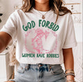 White tee on model with jeans: Model wearing a white God Forbid Women Have Hobbies tee with pink Medusa holding David’s head and green lettering.