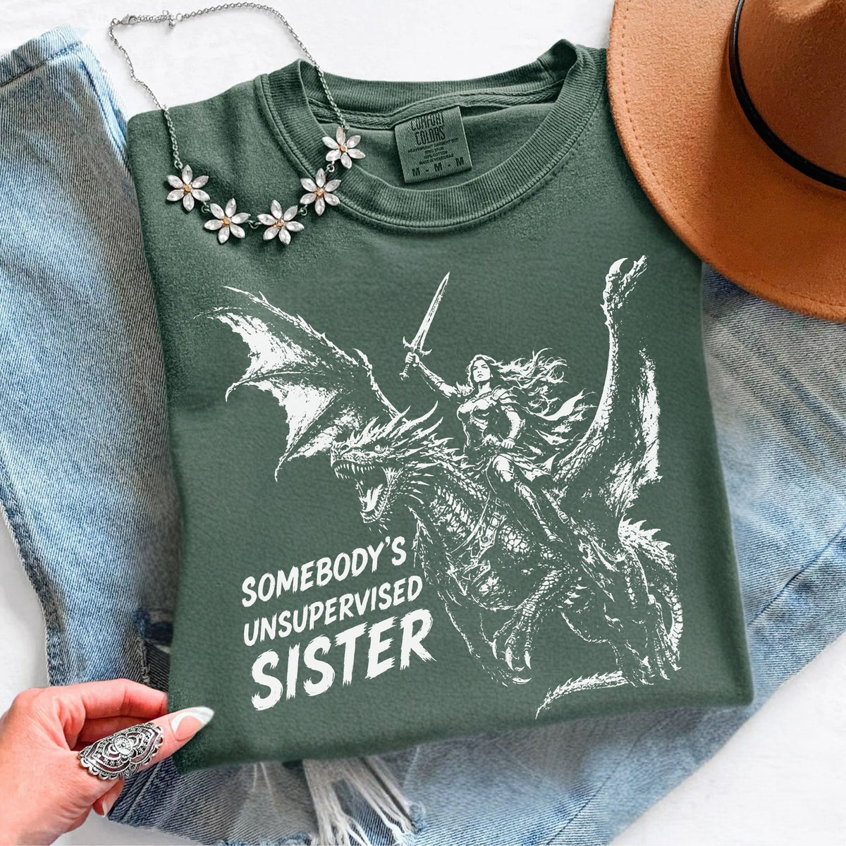 Moss Comfort Colors shirt featuring a female knight riding a dragon with her sword raised and the phrase ‘Somebody’s Unsupervised Sister.