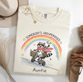 Ivory Somebody’s Unsupervised Auntie T-shirt featuring a raccoon in sunglasses riding a scooter with a cocktail beneath a rainbow.