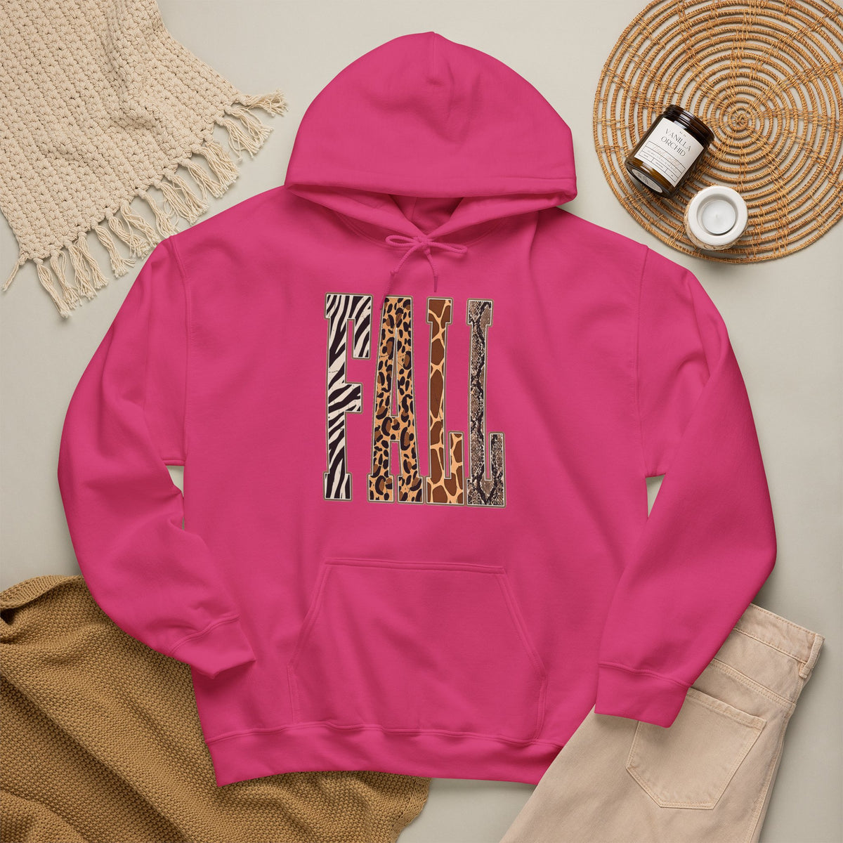A heliconia pullover hoodie laid flat on a light beige surface, featuring large block letters across the chest filled with four animal print patterns: zebra, leopard, giraffe, and cheetah. The hoodie is styled with a tan knit blanket, beige pants, a woven placemat, and small jars in a cozy flat-lay arrangement.