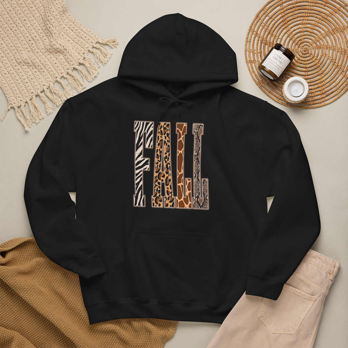 A Black pullover hoodie laid flat on a light beige surface, featuring large block letters across the chest filled with four animal print patterns: zebra, leopard, giraffe, and cheetah. The hoodie is styled with a tan knit blanket, beige pants, a woven placemat, and small jars in a cozy flat-lay arrangement.